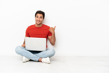 Wall Mural - Caucasian handsome man with a laptop sitting on the floor pointing to the side to present a product