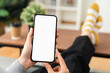 Hands holding smartphone with mockup of blank screen on sofa in living room.