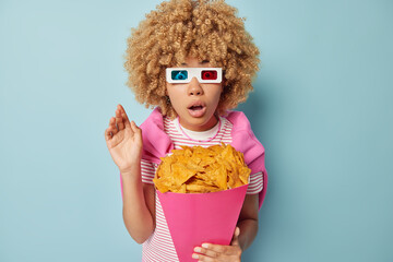 Wall Mural - Impressed shocked curly haired woman wears cinema eyeglasses watches horror film holds paper bucket full of crisps eats delicious snack reacts on something breathtaking isolated over blue background.