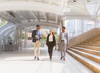 businessman and businesswoman walking with happiness outside building in city with copy space. black man in grey suit, executive man in blue suit and tie holding tablet, caucasian woman in black suit