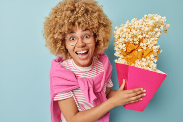 Positive young woman holds paper bucket with spilled popcorn and crisps has fun in cinema foolishes around wears spectacles striped t shirt and pink pullover tied over shoulders poses over blue wall