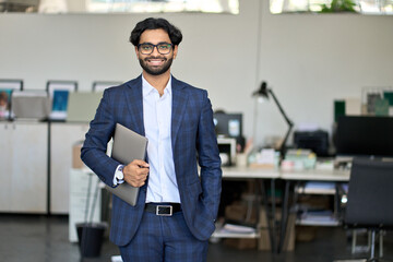 Smiling elegant young indian business man professional manager, arab company employee, eastern businessman executive financial banker wearing suit standing in modern office holding laptop, portrait.