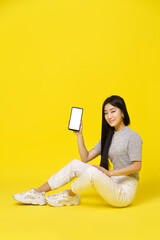 Charming asian young girl holding smartphone sitting on the floor showing a black screen isolated on yellow background. Great offer. Product placement. Mobile App Advertisement. Copy space