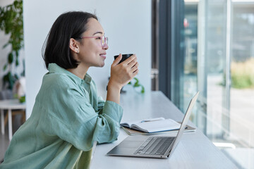 Happy dreamy woman copywriter works on laptop enjoys cozy atmosphere in cafeteria starts day with coffee looks through window admires nice view dressed in casual clothes. female student studies online