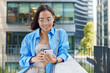 Outdoor shot of beautiful Asian woman with short dark hair uses smartphone device drinks takeaway coffee scrolls social networks wears big transparent eyeglasses blue shirt poses over office building