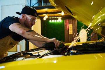 Young repairman bending over engine of car while using electric grinder for repairing details of motor during work in garage