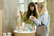 © pressmaster - Blond pretty girl showing body scrub to her friend with dark long wavy hair while choosing bodycare products in cosmetic supermarket