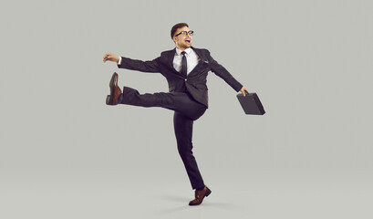 Businessman with crazy facial expression walking strangely, isolated on gray background. Full length of young man in suit with briefcase behaves strangely, takes wide step and shouts something.