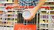Close-up of a shopping trolley in a supermarket and a male buyer puts canned food into it