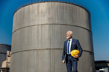 A rich senior businessman walks at his refinery and checks on works. In the background is a tank with oil.