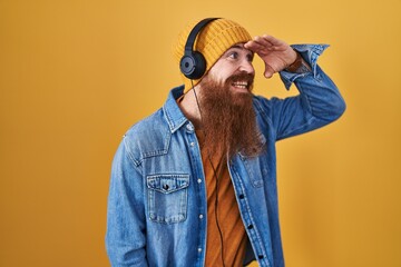 Poster - Caucasian man with long beard listening to music using headphones very happy and smiling looking far away with hand over head. searching concept.