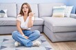 © Krakenimages.com - Young caucasian woman sitting on the floor at the living room surprised pointing with hand finger to the side, open mouth amazed expression.