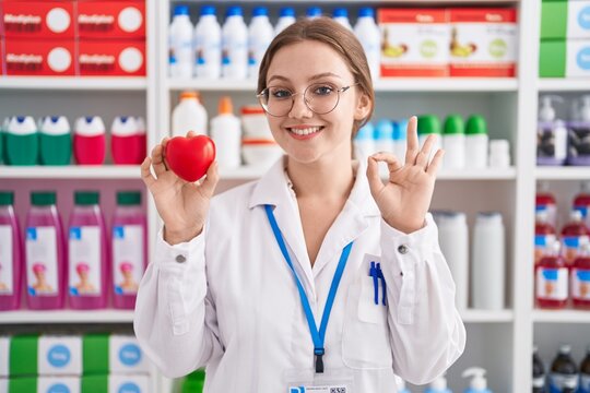 Young caucasian woman working at pharmacy drugstore holding red heart doing ok sign with fingers, smiling friendly gesturing excellent symbol