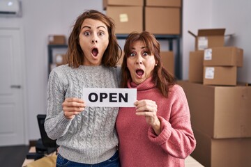 Wall Mural - Two women working at small business ecommerce holding open banner afraid and shocked with surprise and amazed expression, fear and excited face.