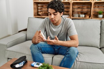 Wall Mural - Young hispanic diabetic man measuring glucose sitting on the sofa at home.