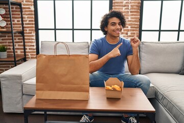 Canvas Print - Hispanic man with curly hair eating chicken wings smiling and looking at the camera pointing with two hands and fingers to the side.