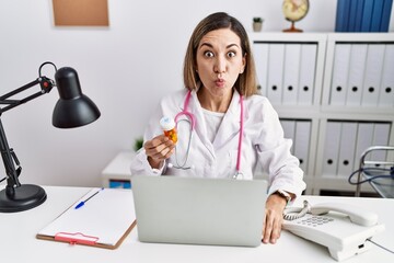 Canvas Print - Young hispanic woman wearing doctor uniform holding pills at the clinic puffing cheeks with funny face. mouth inflated with air, crazy expression.