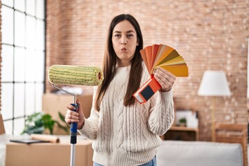 Poster - Young brunette woman holding roller painter and paint samples puffing cheeks with funny face. mouth inflated with air, catching air.