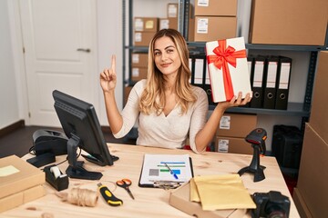 Poster - Young blonde woman working at small business ecommerce holding gift smiling with an idea or question pointing finger with happy face, number one