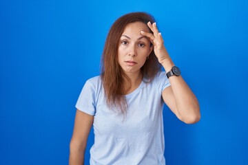 Wall Mural - Brunette woman standing over blue background worried and stressed about a problem with hand on forehead, nervous and anxious for crisis