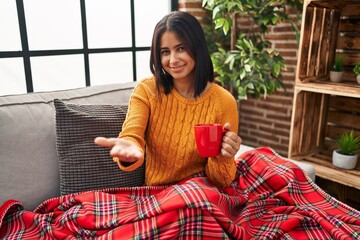 Sticker - Young hispanic woman sitting on the sofa drinking a coffee at home smiling cheerful offering palm hand giving assistance and acceptance.