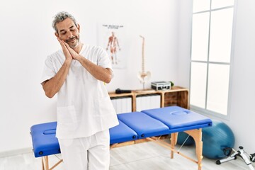 Poster - Middle age hispanic therapist man working at pain recovery clinic sleeping tired dreaming and posing with hands together while smiling with closed eyes.