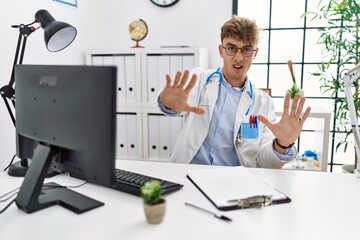 Wall Mural - Young caucasian doctor man working at the clinic afraid and terrified with fear expression stop gesture with hands, shouting in shock. panic concept.