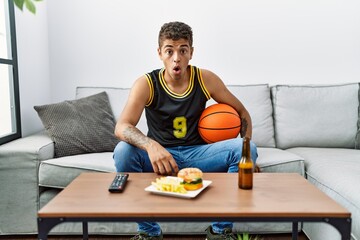 Wall Mural - Young handsome hispanic man holding basketball ball cheering tv game scared and amazed with open mouth for surprise, disbelief face