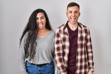 Poster - Young hispanic couple standing over white background sticking tongue out happy with funny expression. emotion concept.