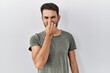 Young hispanic man with beard wearing casual t shirt over white background smelling something stinky and disgusting, intolerable smell, holding breath with fingers on nose. bad smell