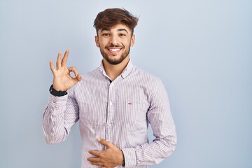 Poster - Arab man with beard standing over blue background smiling positive doing ok sign with hand and fingers. successful expression.