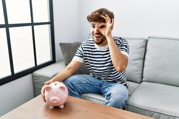 Poster - Young arab man holding piggy bank smiling happy doing ok sign with hand on eye looking through fingers