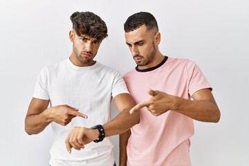 Canvas Print - Young gay couple standing over isolated background in hurry pointing to watch time, impatience, upset and angry for deadline delay