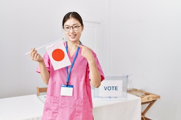 Sticker - Young asian nurse woman at political campaign election holding japan flag smiling happy pointing with hand and finger