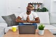 Young african man using laptop at home begging and praying with hands together with hope expression on face very emotional and worried. begging.