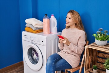 Wall Mural - Young woman using smartphone waiting for washing machine at laundry room