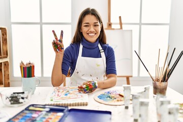 Canvas Print - Young brunette woman at art studio with painted hands showing and pointing up with fingers number two while smiling confident and happy.