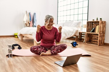 Poster - Middle age woman with grey hair training at home looking at exercise video on laptop screaming proud, celebrating victory and success very excited with raised arms