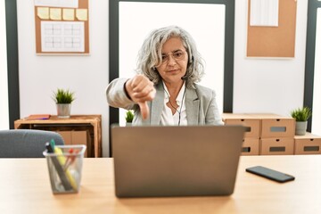 Sticker - Middle age businesswoman sitting on desk working using laptop at office looking unhappy and angry showing rejection and negative with thumbs down gesture. bad expression.