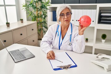 Poster - Middle age woman with grey hair wearing doctor uniform holding balloon thinking attitude and sober expression looking self confident