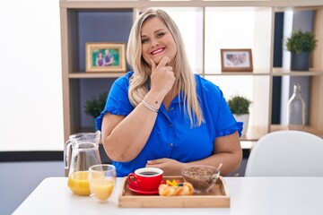 Poster - Caucasian plus size woman eating breakfast at home looking confident at the camera smiling with crossed arms and hand raised on chin. thinking positive.