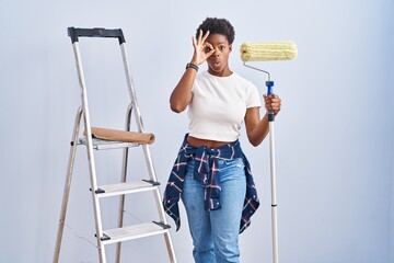 Wall Mural - African american woman holding roller painter doing ok gesture shocked with surprised face, eye looking through fingers. unbelieving expression.