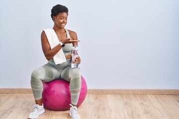 Poster - African american woman wearing sportswear sitting on pilates ball pointing aside with hands open palms showing copy space, presenting advertisement smiling excited happy