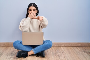 Sticker - Young woman using laptop sitting on the floor at home laughing at you, pointing finger to the camera with hand over mouth, shame expression