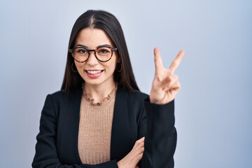 Canvas Print - Young brunette woman standing over blue background smiling with happy face winking at the camera doing victory sign. number two.