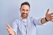 © Krakenimages.com - Middle age caucasian man standing over blue background looking at the camera smiling with open arms for hug. cheerful expression embracing happiness.