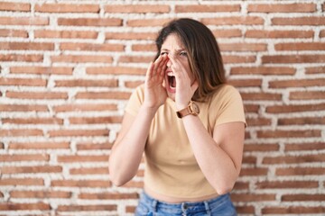 Sticker - Young brunette woman standing over bricks wall shouting angry out loud with hands over mouth