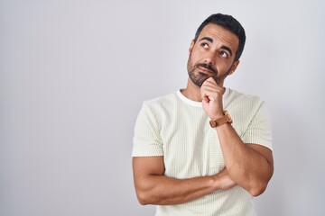 Canvas Print - Hispanic man with beard standing over isolated background with hand on chin thinking about question, pensive expression. smiling with thoughtful face. doubt concept.