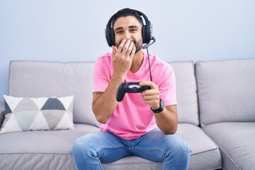 Wall Mural - Hispanic young man playing video game holding controller sitting on the sofa laughing and embarrassed giggle covering mouth with hands, gossip and scandal concept