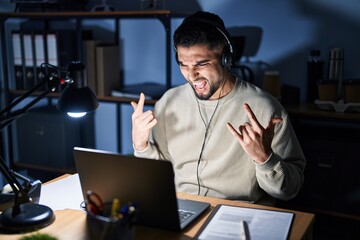 Wall Mural - Young handsome man working using computer laptop at night shouting with crazy expression doing rock symbol with hands up. music star. heavy concept.